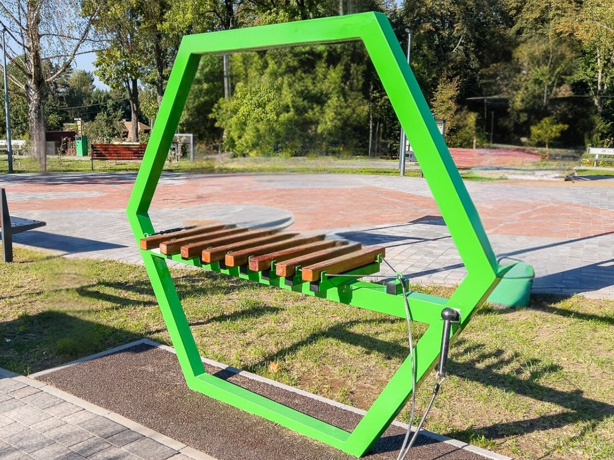 Zielona stalowa rama ksylofonu z&nbsp;zawieszonym rzędem drewnianych płytek, ustawiona na placu zabaw