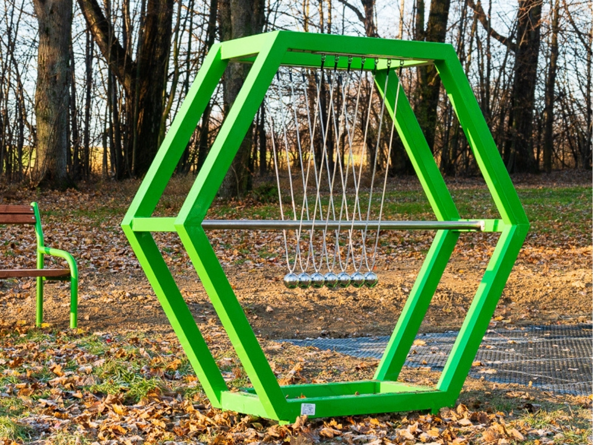 The powder-coated steel frame of the Newton's cradle with a row of balls hanging on cables, the playground in the background