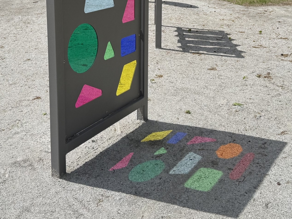 Grafitowy panel sensoryczny w&nbsp;słońcu, a&nbsp;na jasnej nawierzchni w&nbsp;jego cieniu kolorowe figury: zielony owal, żółty równoległobok, różowy trójkąt