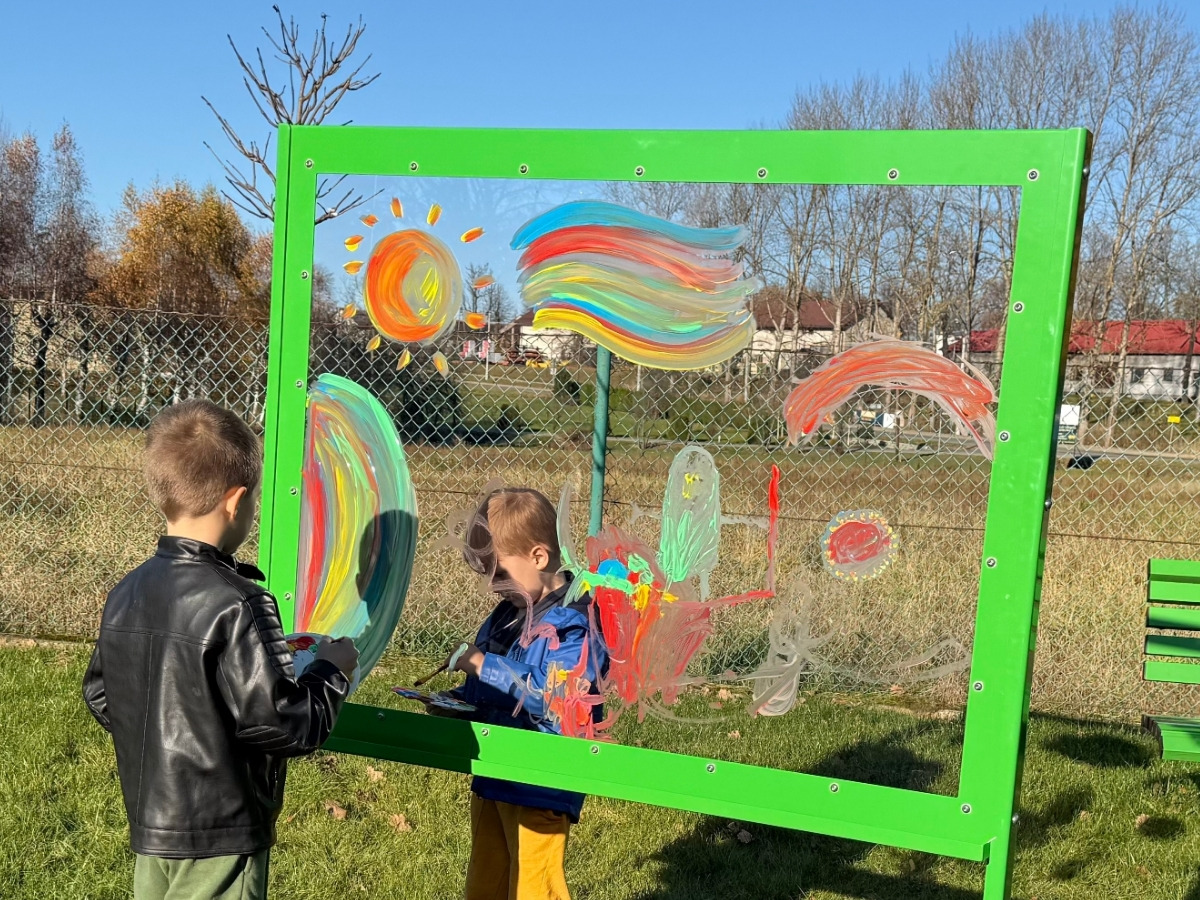Two children painting at the same time on opposite sides of the transparent panel, their pictures overlapping, their faces visible through it