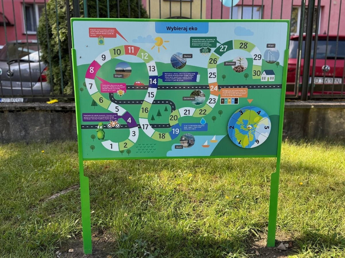 Tablica z&nbsp;grą planszową na stalowym słupku osadzonym w&nbsp;trawniku, obok stoi dziecko