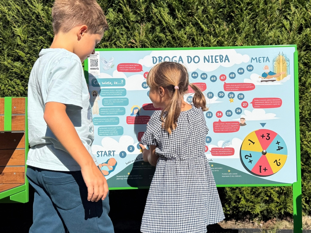 Tablica z&nbsp;grą planszową na stalowym słupku osadzonym w&nbsp;trawniku, obok stoi dziecko