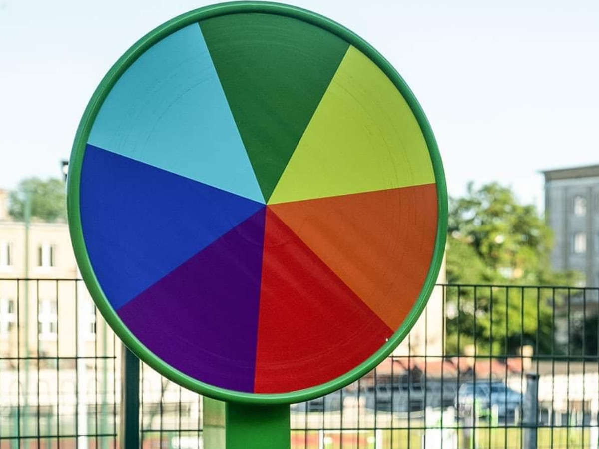 Koło edukacyjne w&nbsp;zielonej ramie z&nbsp;tarczą podzieloną na siedem kolorowych pól, w&nbsp;tle ogrodzenie i&nbsp;budynki