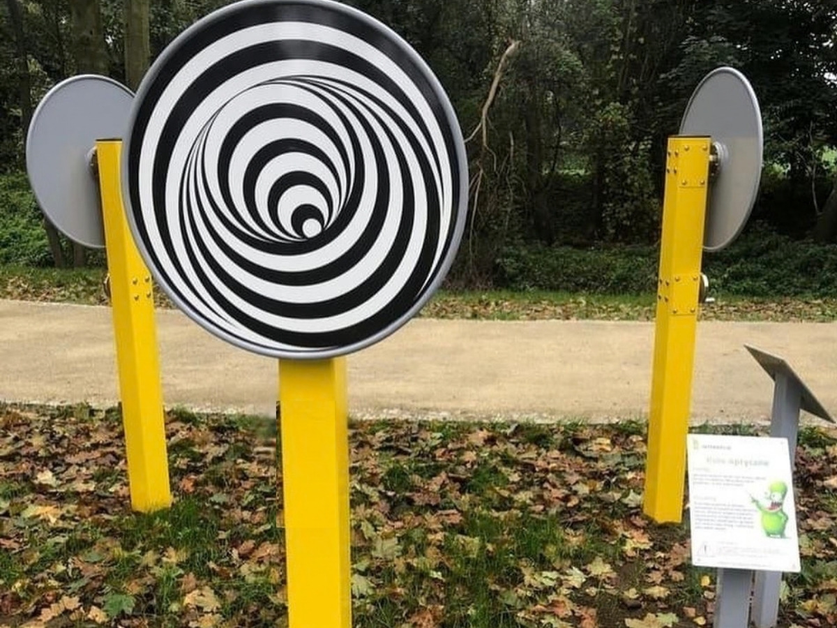 Koło optyczne z&nbsp;czarno-białą spiralą na żółtym słupku, obok dwa kolejne koła na żółtych słupkach, w&nbsp;tle alejka i&nbsp;drzewa