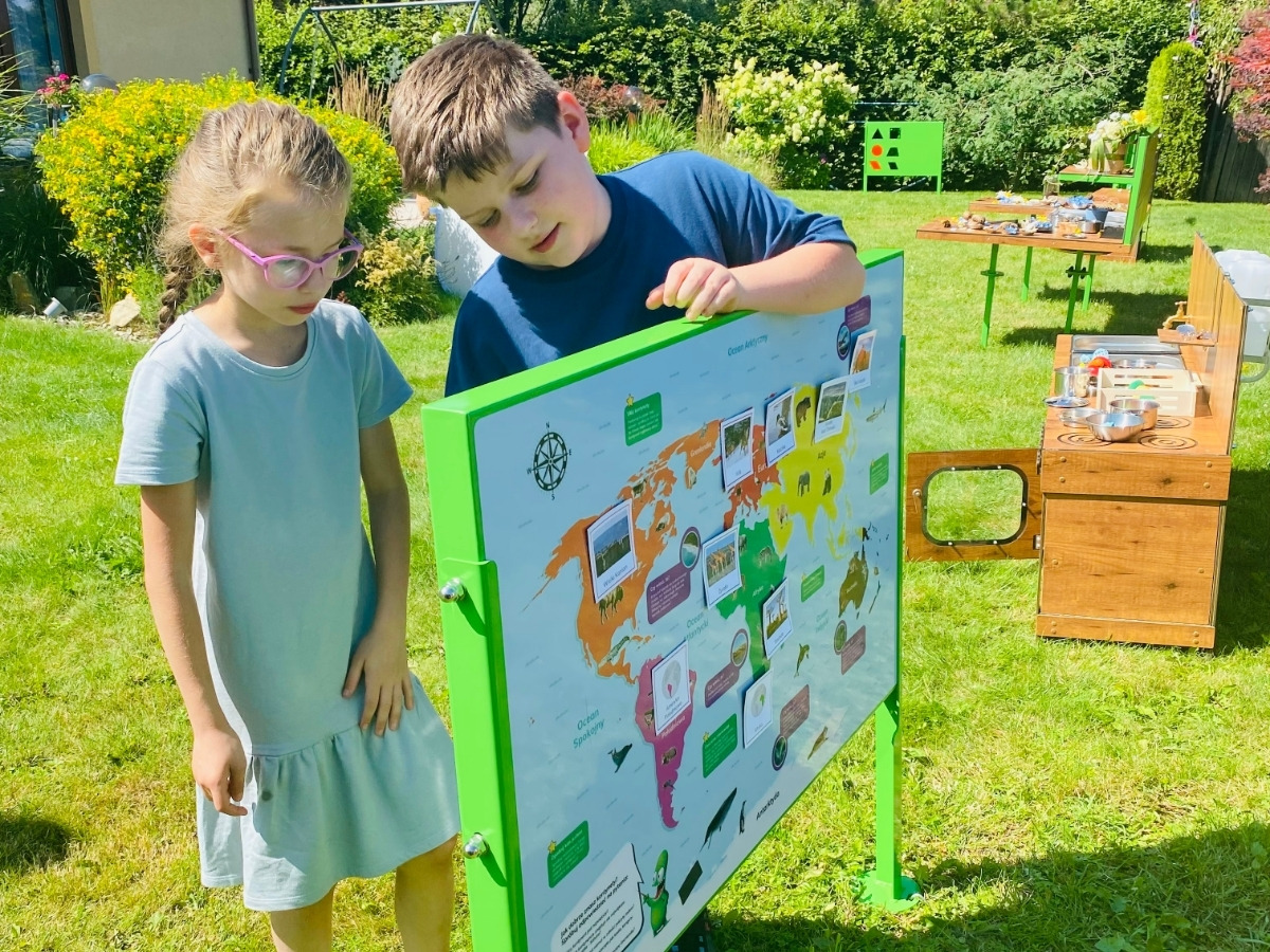 Dwoje dzieci przy tablicy edukacyjnej z&nbsp;mapą świata na trawniku, chłopiec oparty o&nbsp;bok tablicy