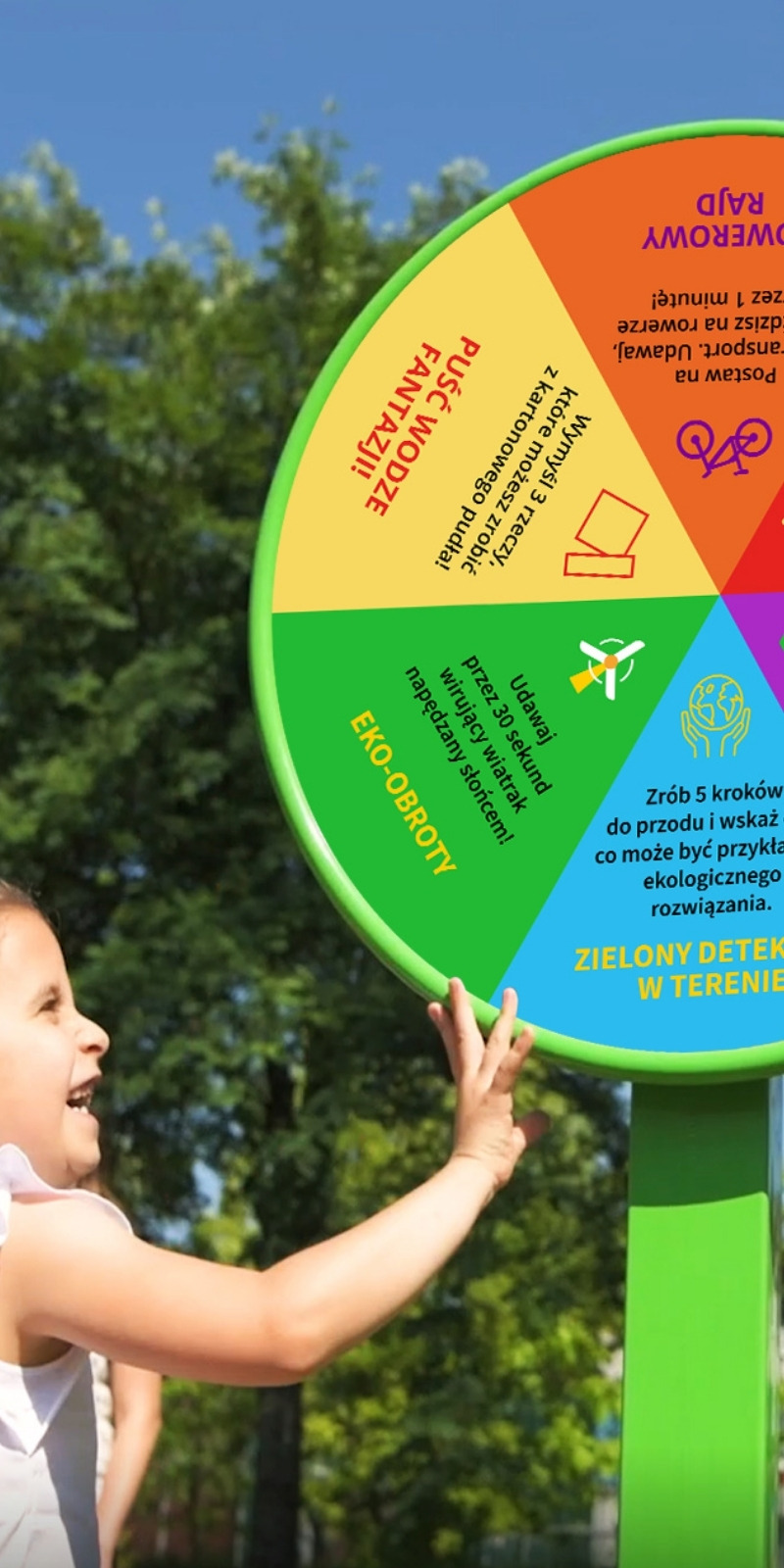 Ekologiczny plac zabaw: dziecko obraca duże kolorowe koło z polami „Eko-obroty” i „Zielony detektyw w terenie”